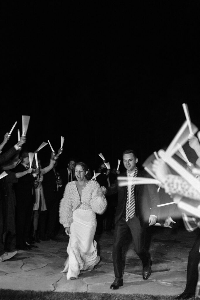 black and white photo of couple during their wedding exit and people cheering with glow wands